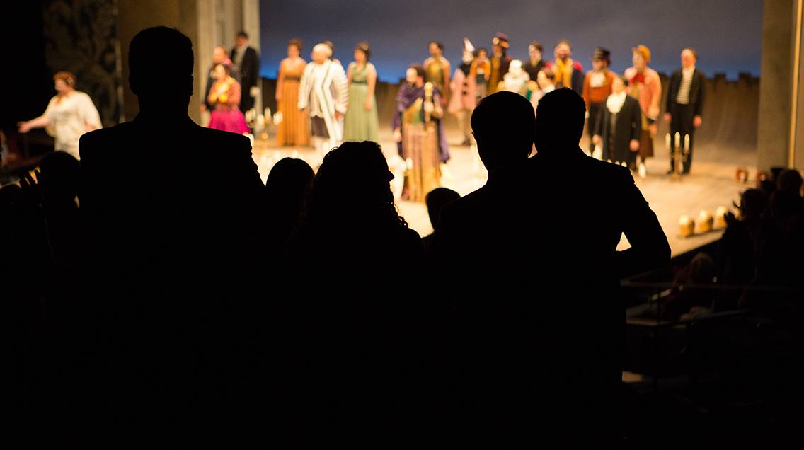 An audience is silhouetted against the stage as members of the cast take their bows.