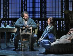 a bearded man in glasses sitting on a chair looks over at his love interest, a girl sitting on a chair with both her hands on her knees
